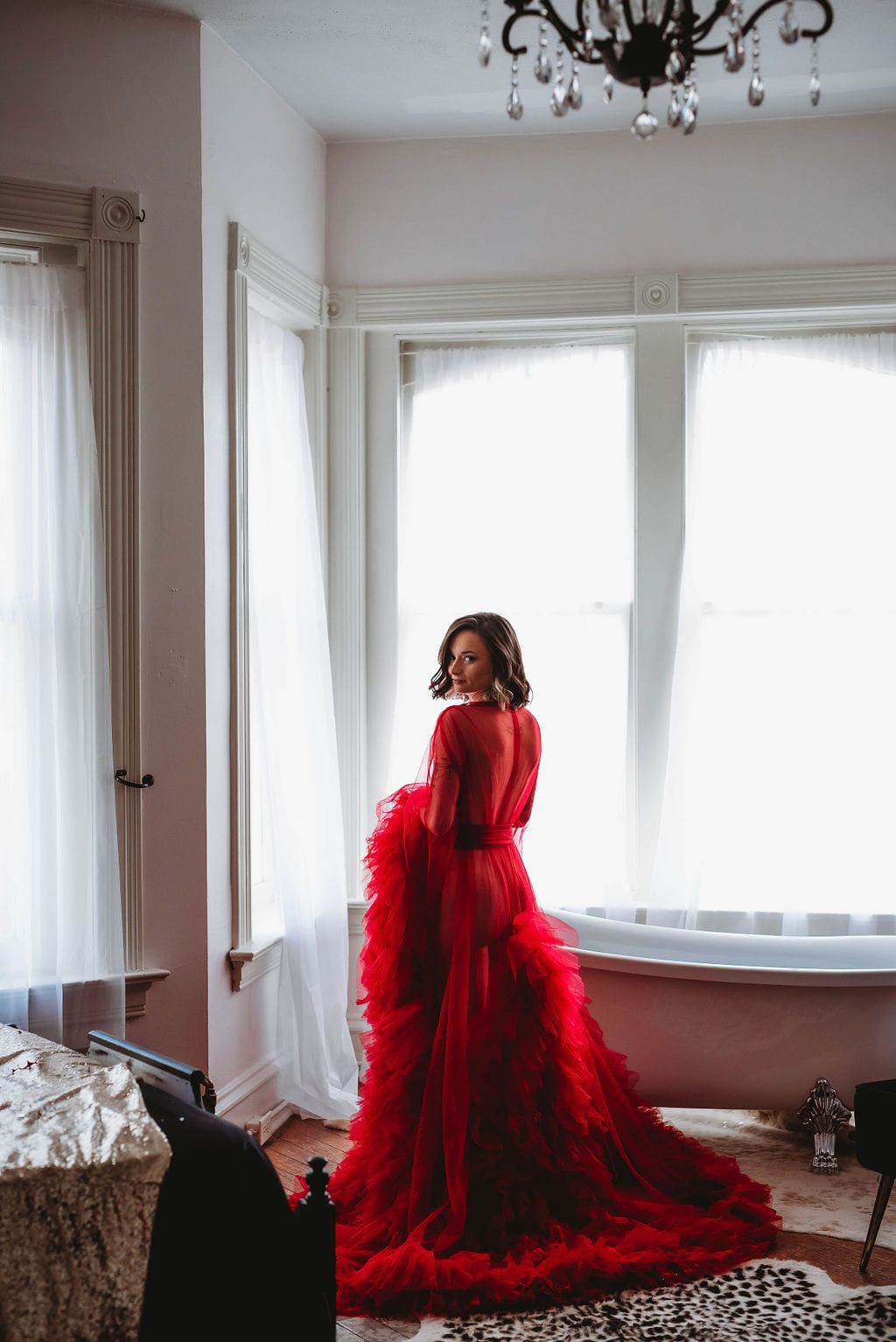 A dramatic flowing red dress with a long train stands in an elegant room with sheer curtains and a chandelier.