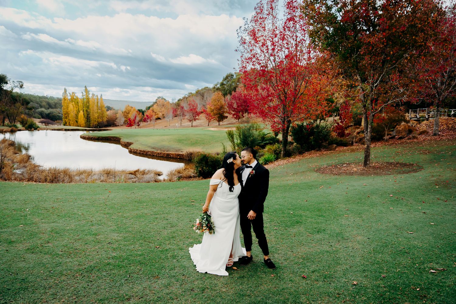 Newlyweds walking on lush grass near a lake surrounded by colorful autumn trees.