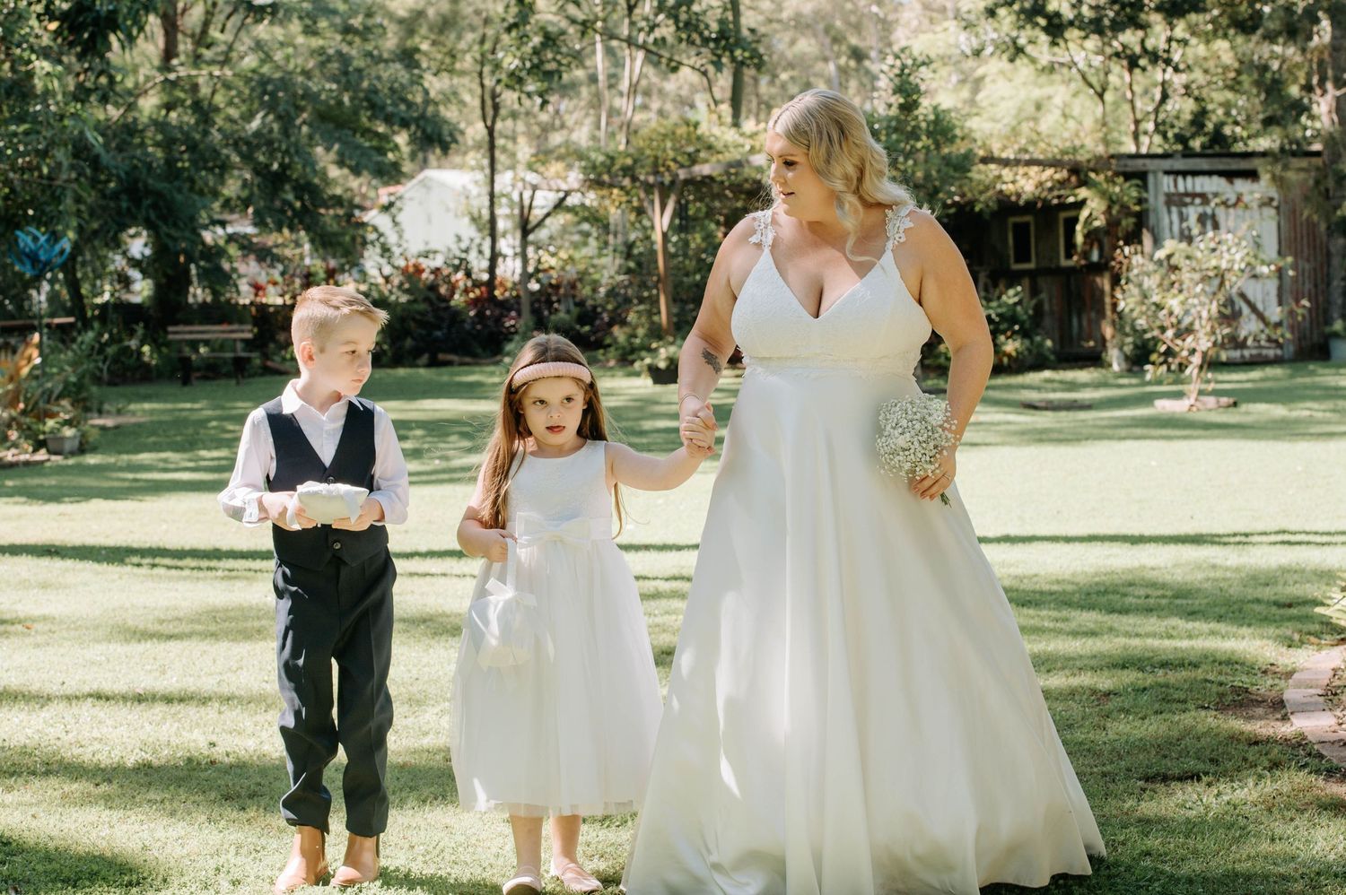 A woman in a wedding dress stands with two children on a sunny lawn during a wedding celebration.