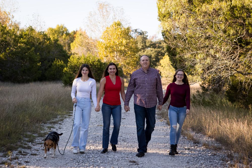 A group walks together with their small dog on a gravel path through tall grass and autumn trees.