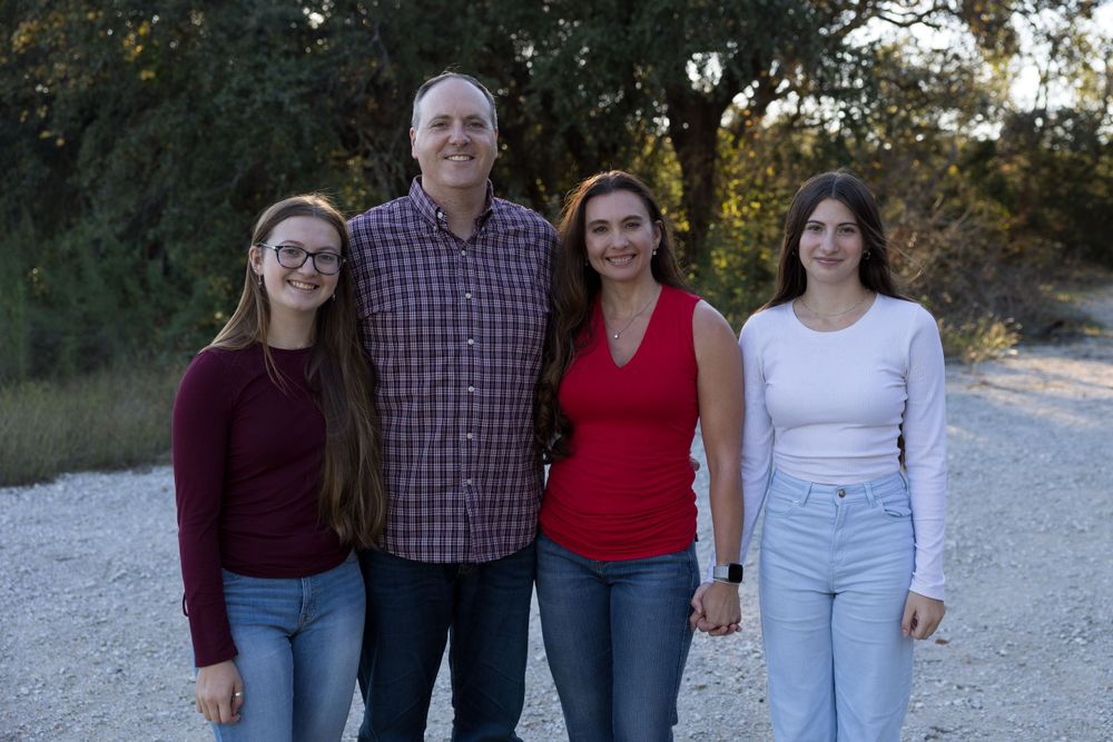 Family of four poses together on gravel path with trees in background.
