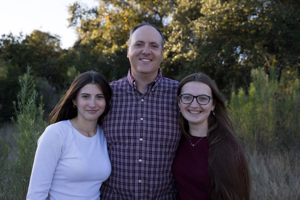 Family of three poses together outdoors in casual attire against natural backdrop.