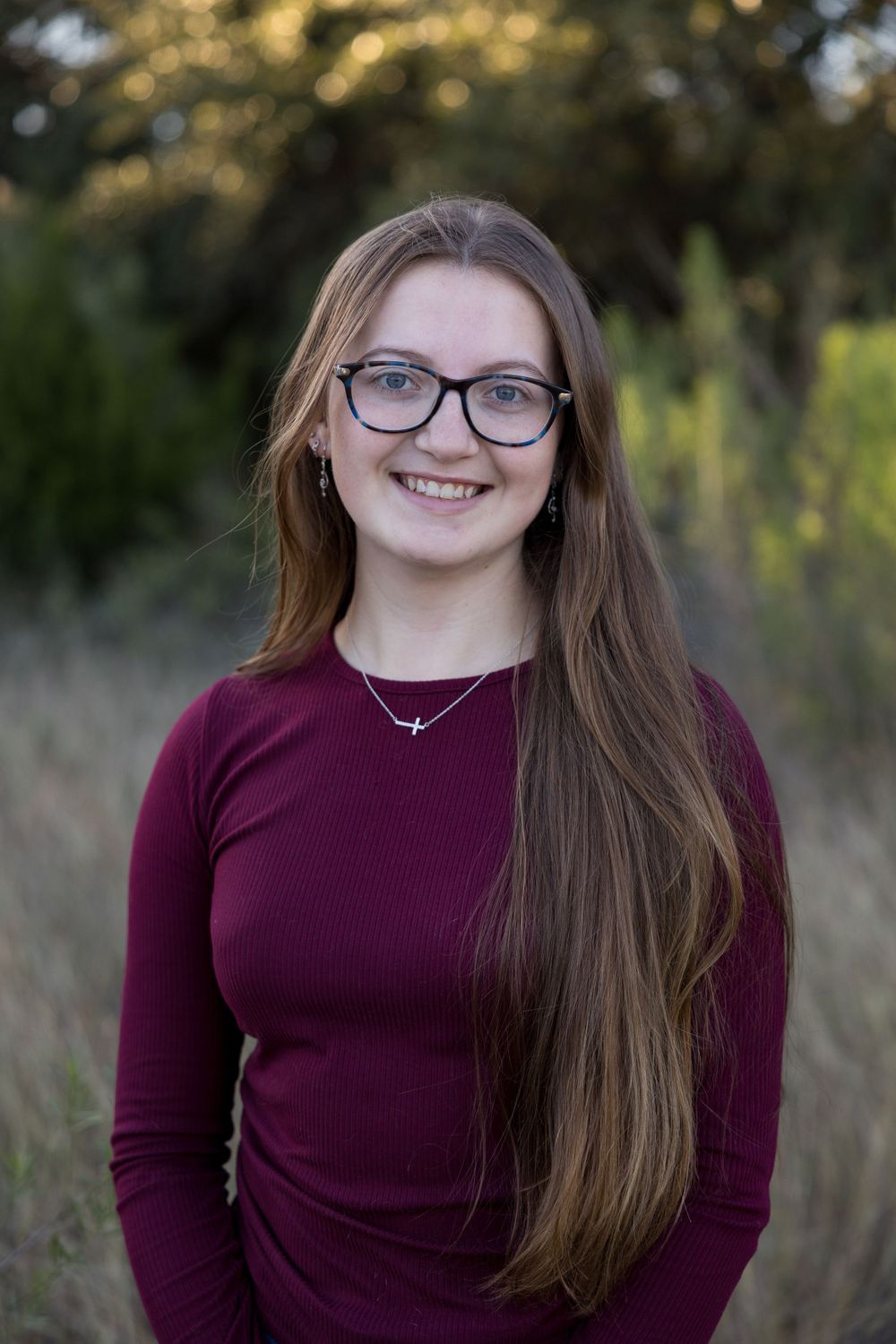 Young woman in burgundy sweater and glasses smiles at camera in outdoor nature setting.