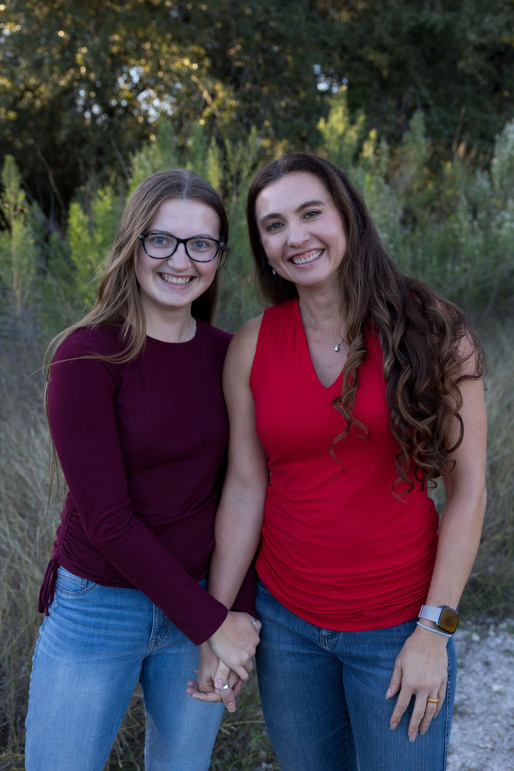 Two women in casual attire smiling while standing together outdoors in a natural setting.