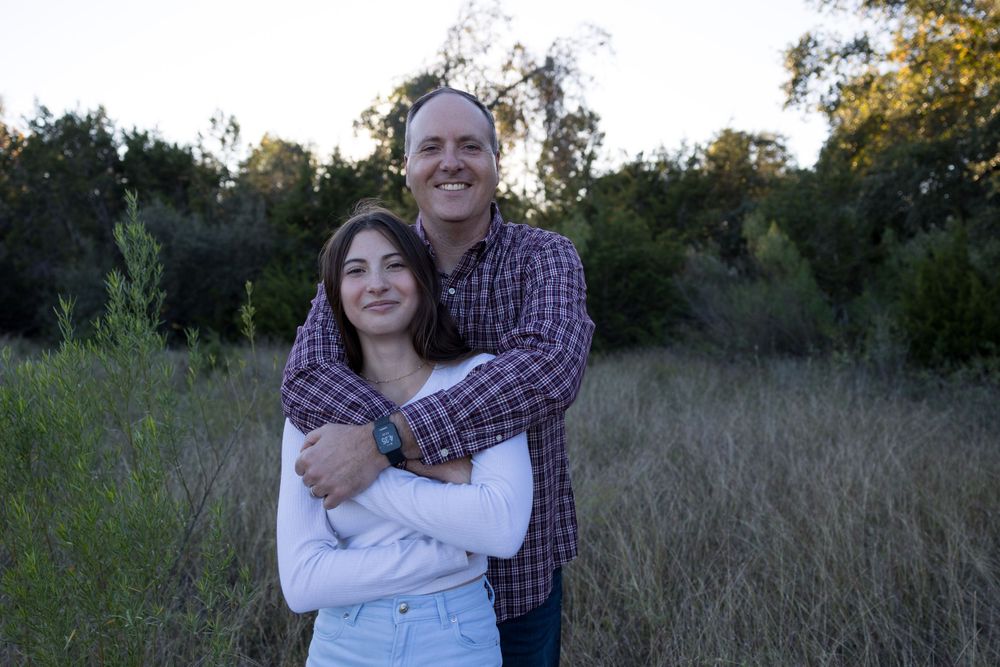 Couple embraces in a natural outdoor setting with tall grass and trees at sunset.