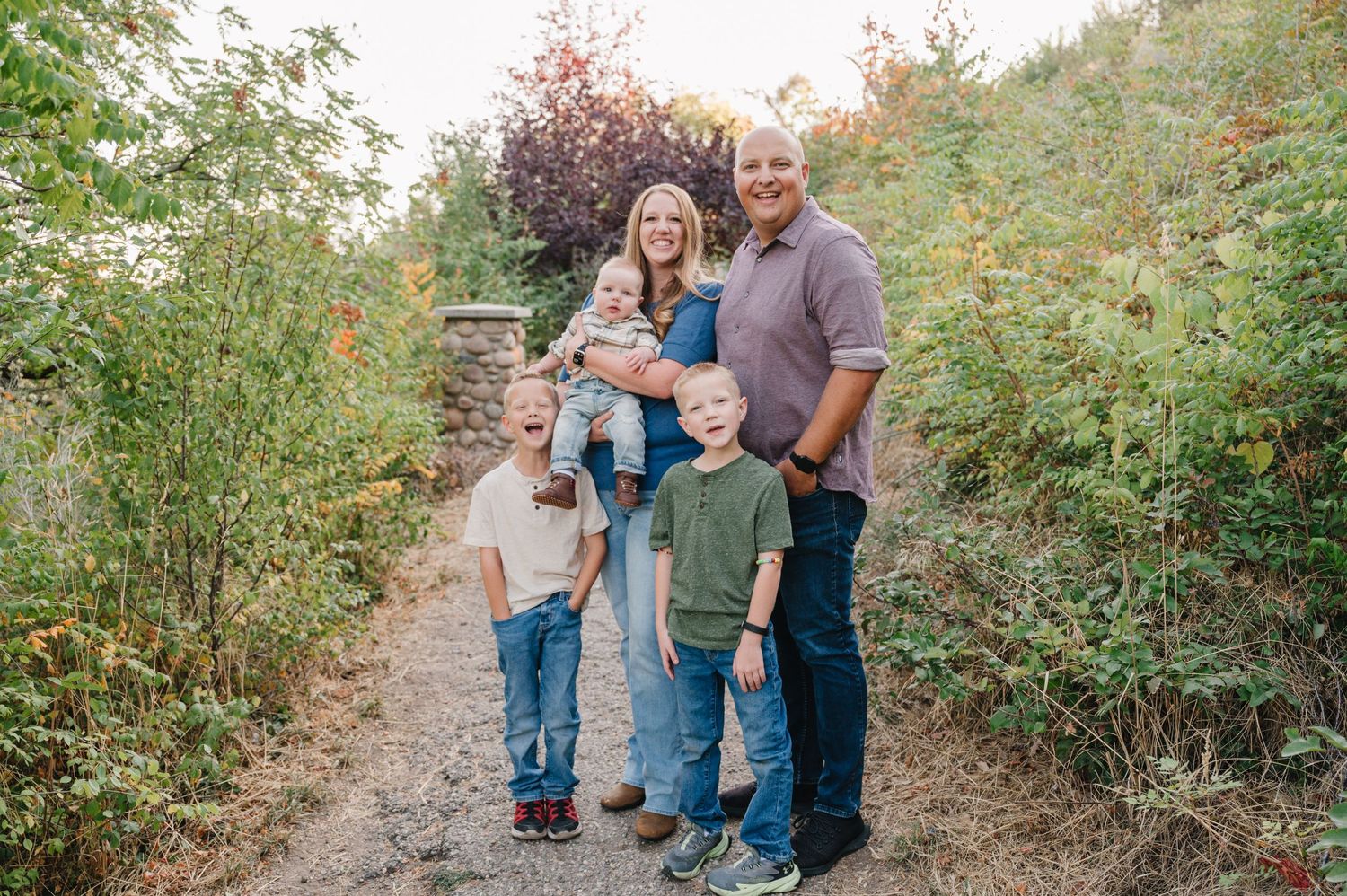 A family stands together on a nature trail surrounded by greenery and autumn foliage on a sunny afternoon.