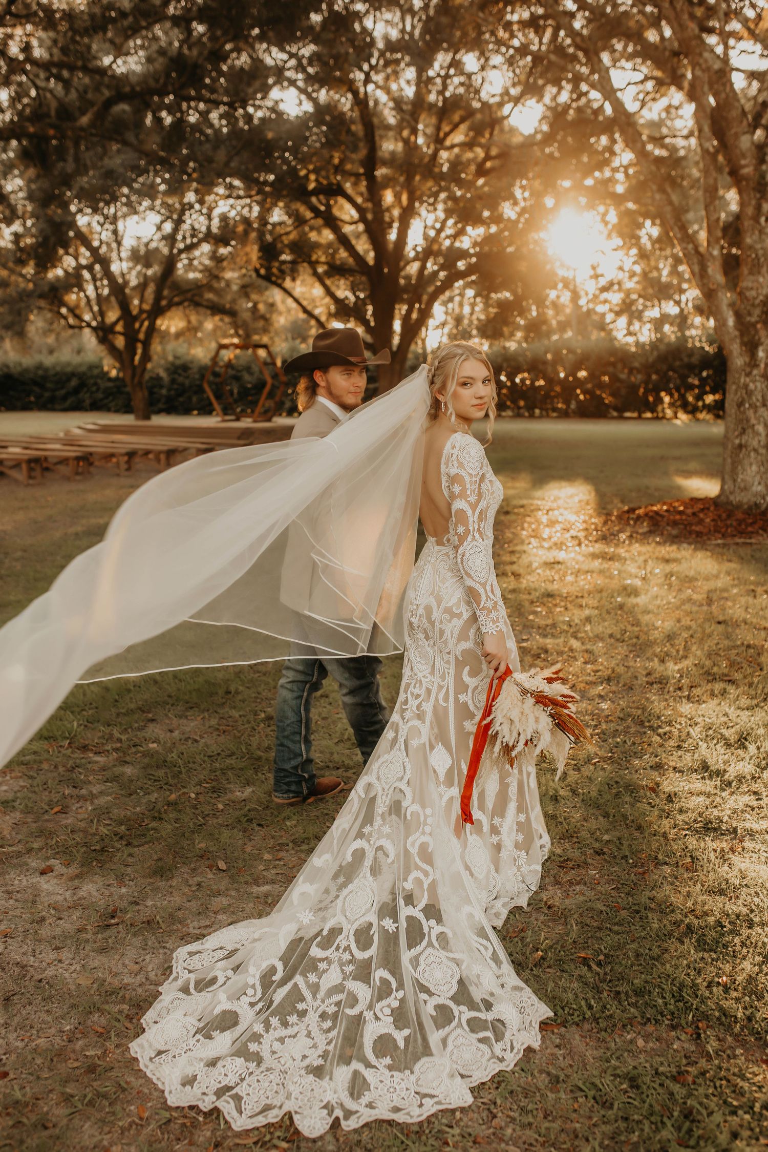 A romantic sunset wedding photo with a flowing veil and intricate lace dress.