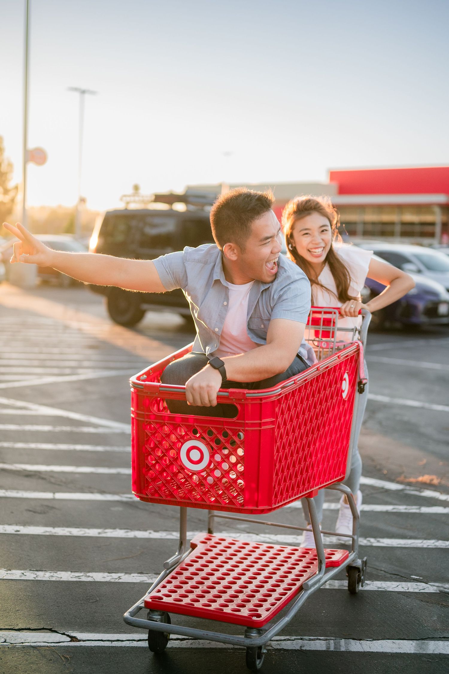 Two people having fun riding in a red Target shopping cart in a parking lot at sunset.