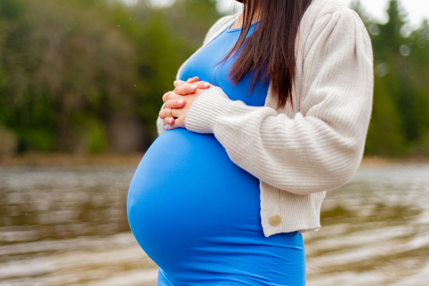 Close up of a pregnant belly in a fitted blue dress and cream cardigan by a lake.