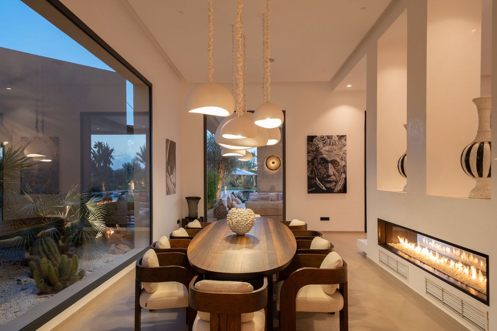 Modern dining room with long wooden table, pendant lights, and linear fireplace beside large window at dusk.
