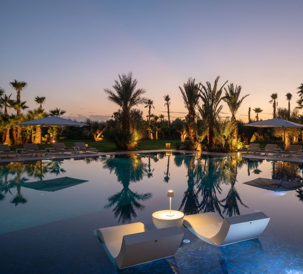 Tranquil resort pool reflecting palm trees and sunset sky with curved lounge seating areas along the water's edge.