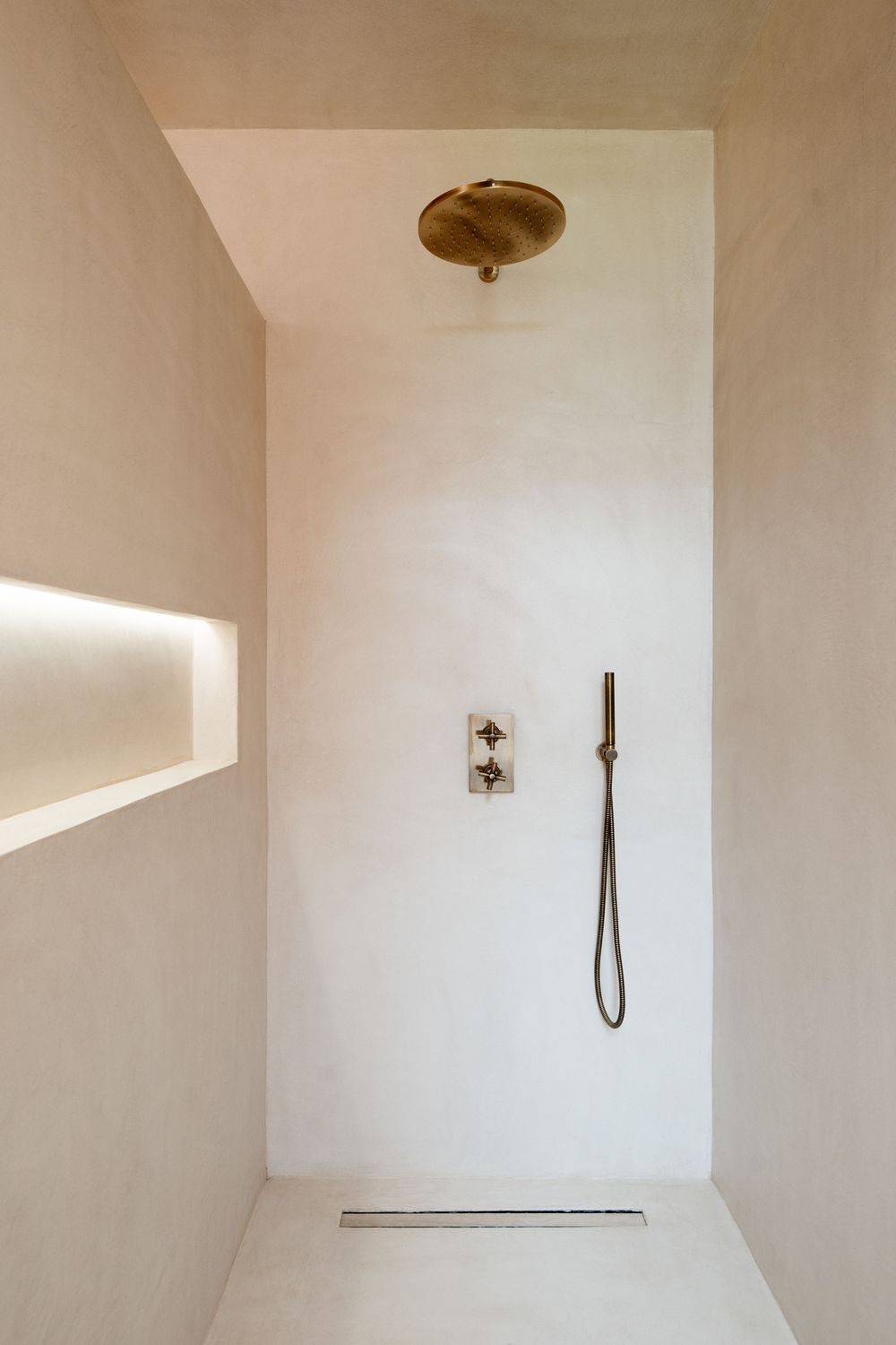 Minimalist white bathroom shower space with brass fixtures and linear drain featuring indirect LED lighting.