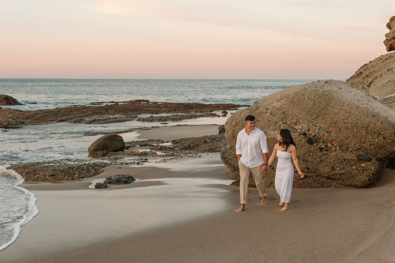 Couple walking hand in hand on beach during sunset near rocky coastline in Laguna Beach