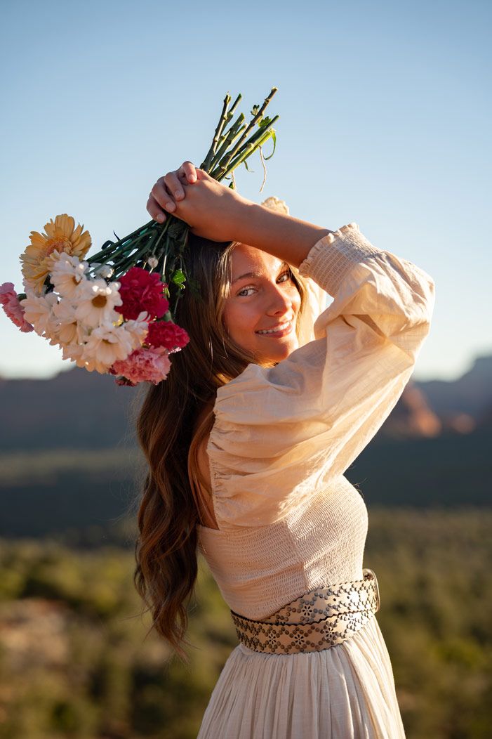 Person in cream dress holds flowers above head against Sedona mountain backdrop at golden hour.