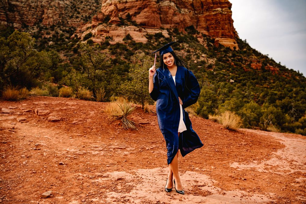 Graduate in blue regalia poses gracefully on Sedona red desert terrain with mountains in background.