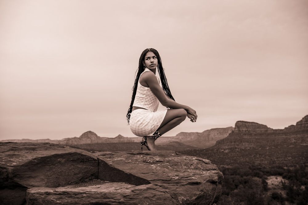 Sepia toned portrait of person in light dress crouching on desert rocks.