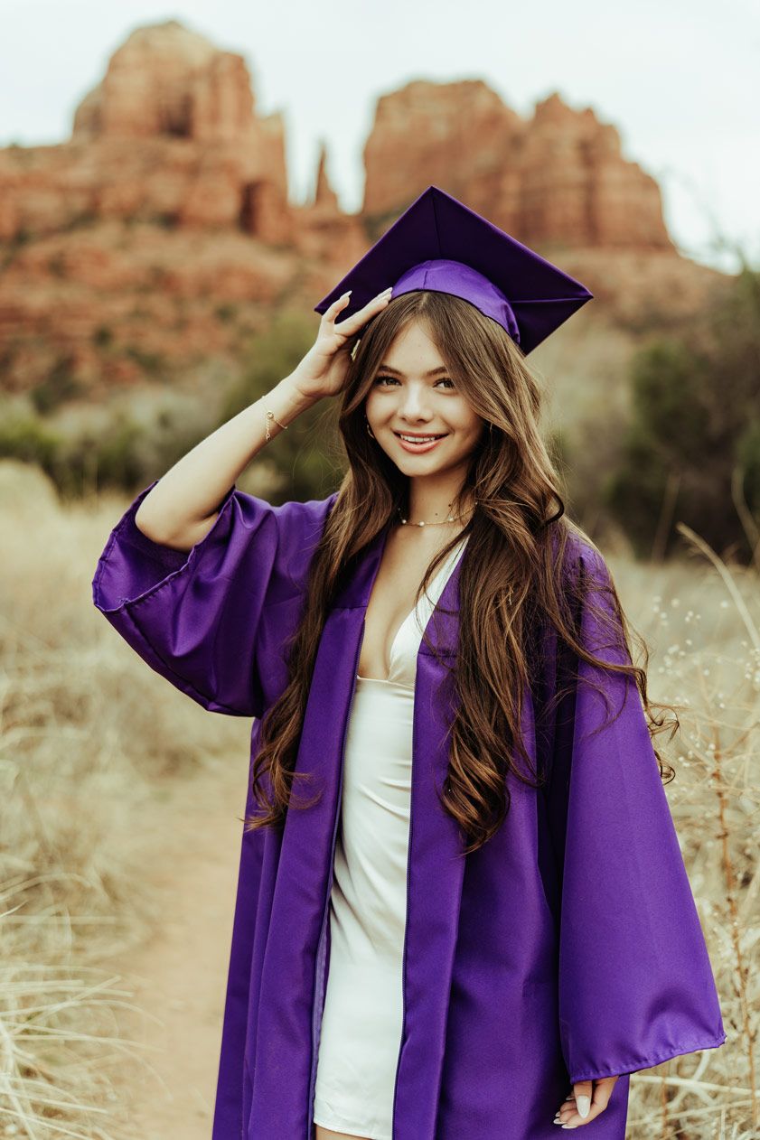 Sedona Red Rock High School raduate in purple cap and gown poses against red rock formations in outdoor desert setting.