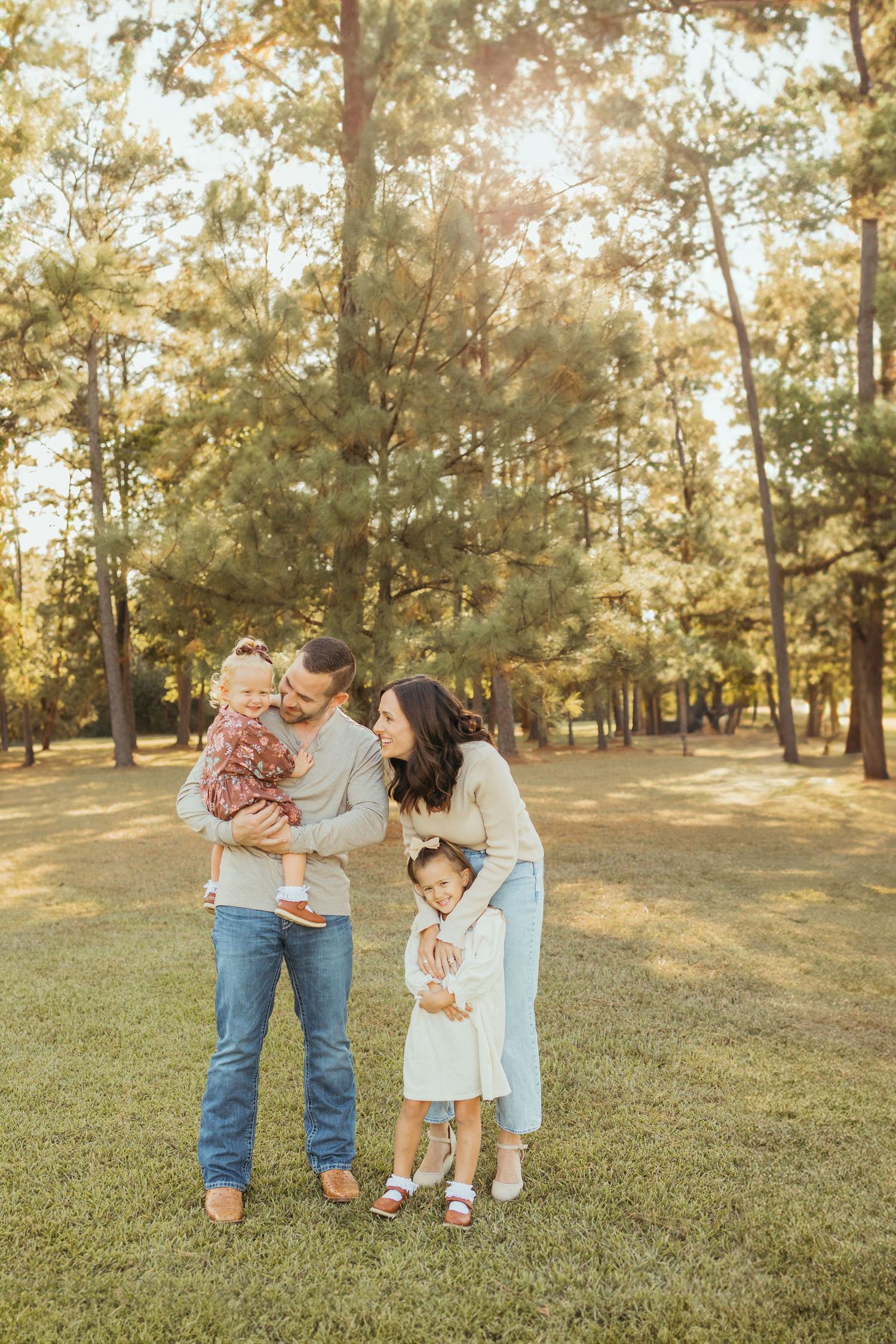 Smiling mother holding toddler while young daughter hugs her leg during outdoor family portrait session.