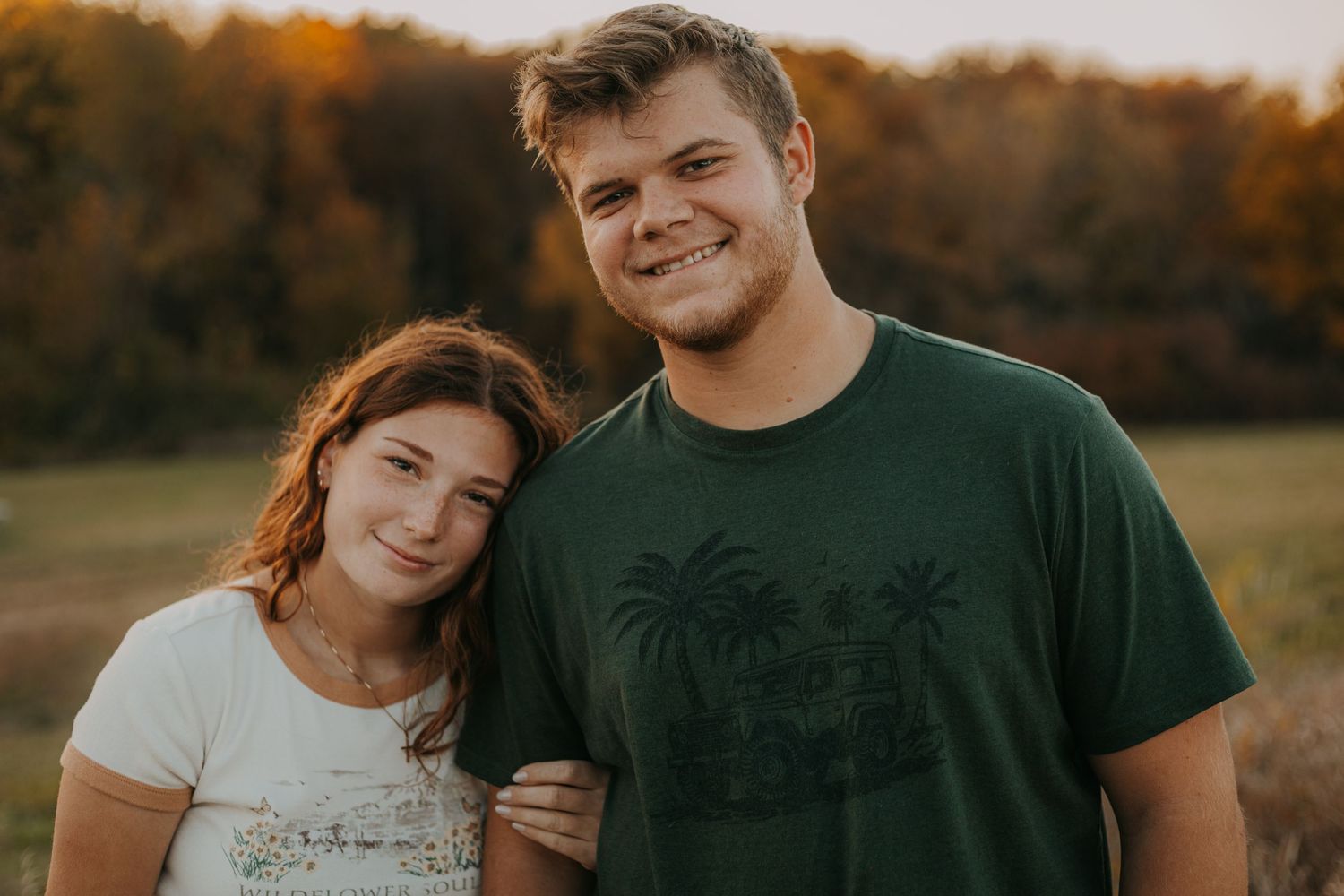 Two people stand close together in a field during golden hour with autumn foliage behind.