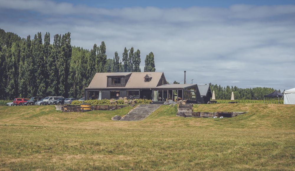 A rustic farmhouse with a dark roof sits on a green meadow surrounded by tall poplar trees under a cloudy sky.