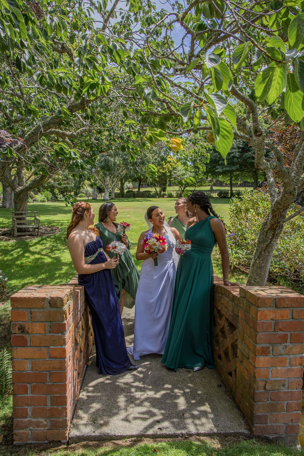 Bride in white gown with three bridesmaids in navy and green dresses holding bouquets in a lush garden setting.