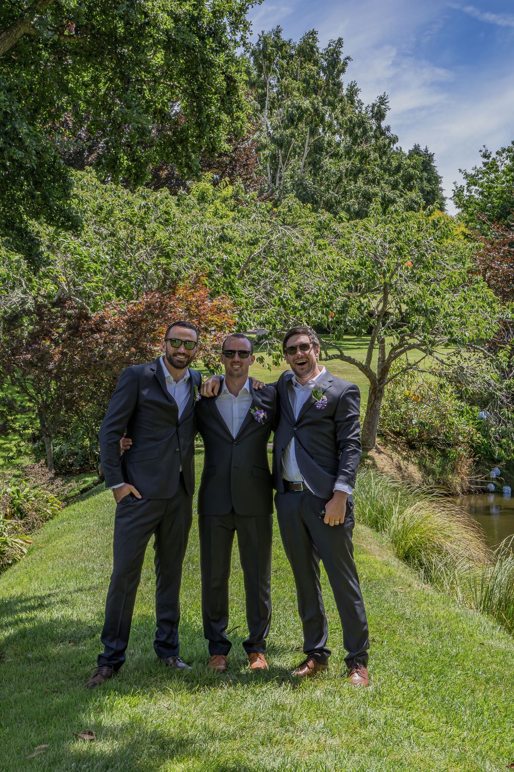 Three men in dark suits posing together outdoors in a lush garden setting on a sunny day.
