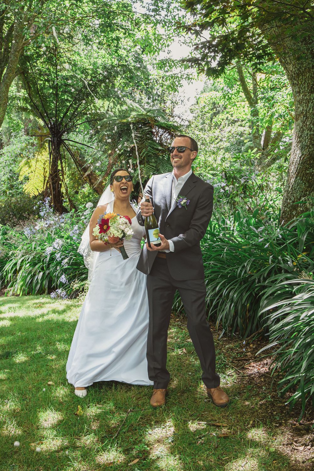 Joyful bride and groom popping champagne in a lush green garden on their wedding day.