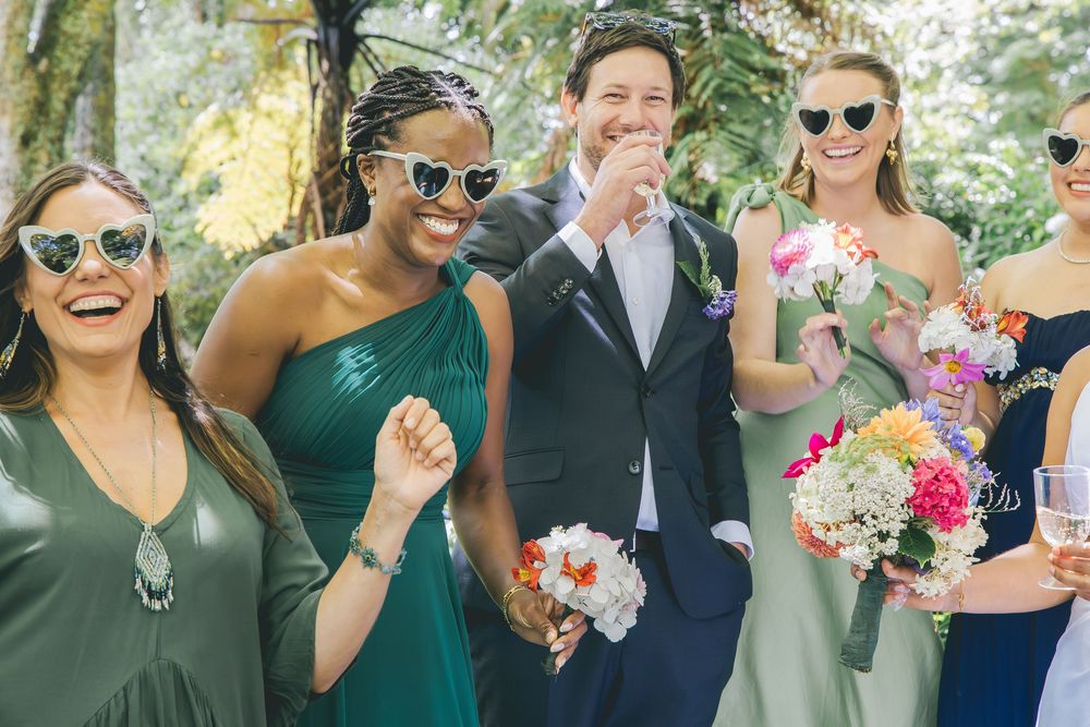 Bridal party in green dresses and heart sunglasses laughing with groom in navy suit outdoors.