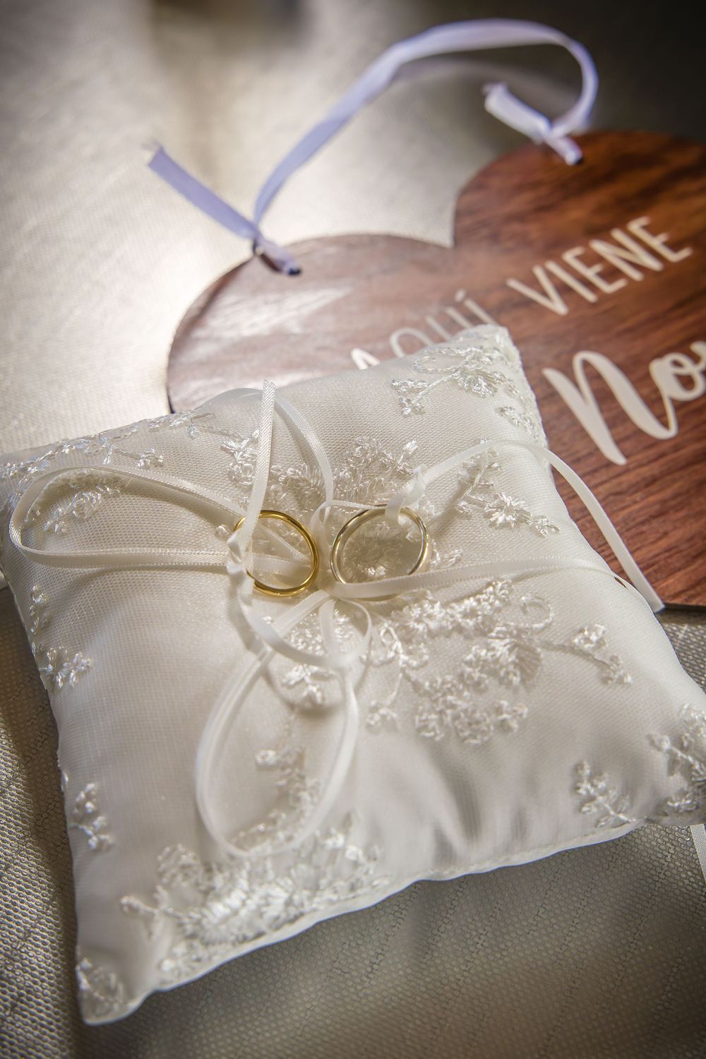 Gold wedding rings rest on a white lace embroidered ring bearer pillow beside a wooden heart sign.