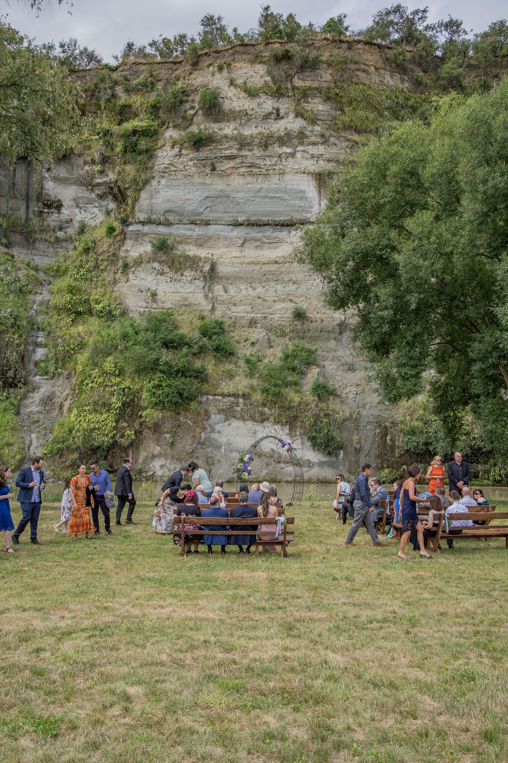 People gather on benches and grass in front of a large white rock cliff face surrounded by lush green trees outdoors.
