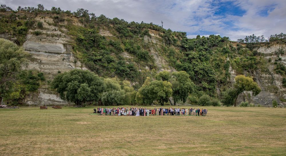 Group of people gathered on green field below dramatic sandstone cliffs with lush trees in New Zealand landscape.