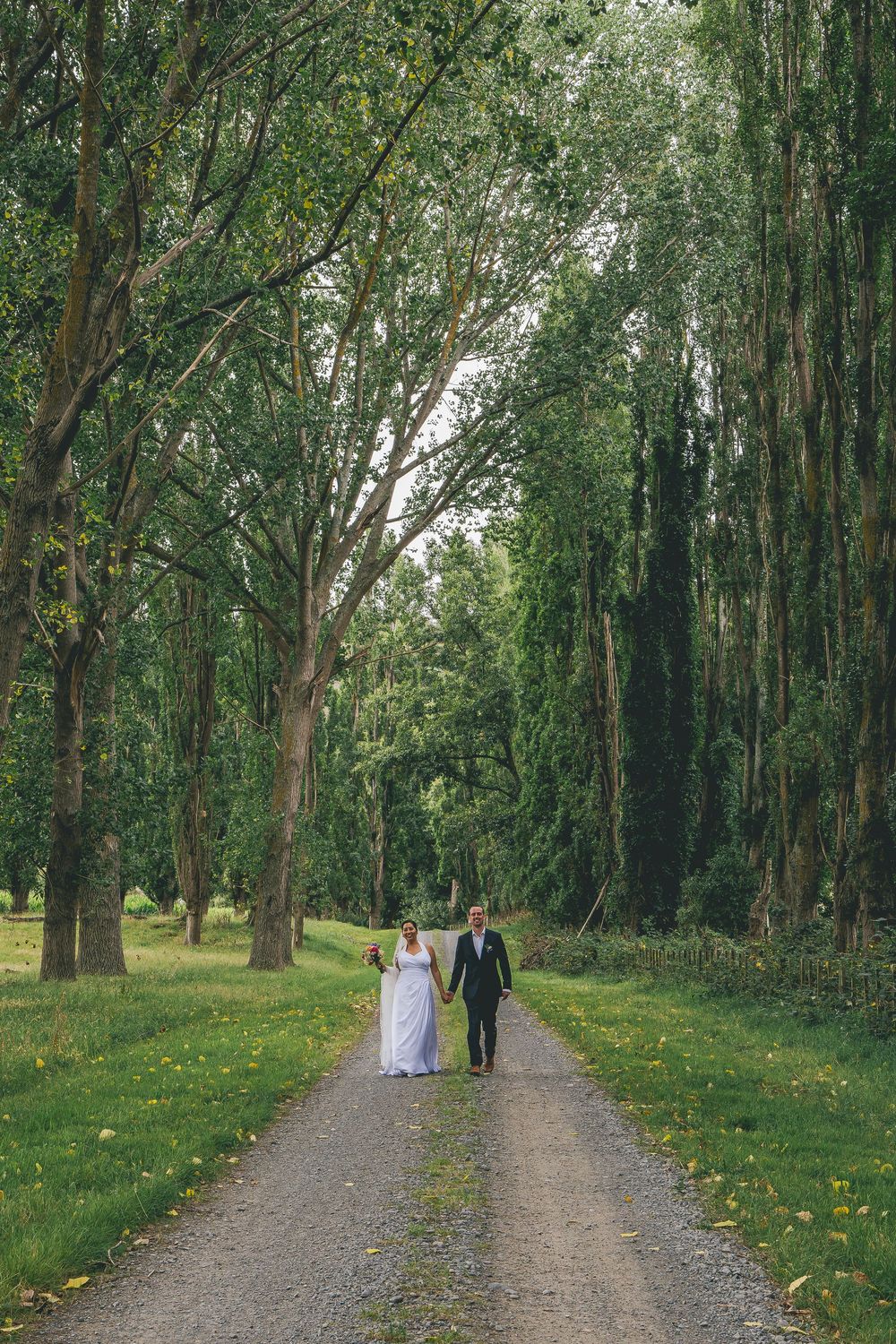 Bride and groom walking hand in hand down a tree-lined gravel path surrounded by lush green foliage.