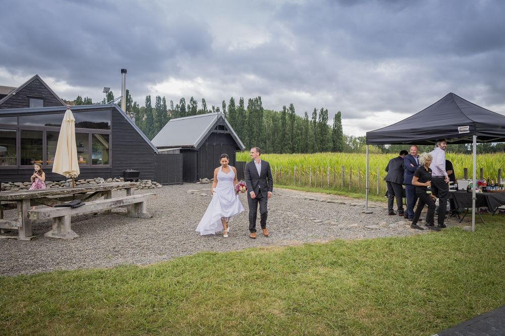 Bride and groom walking at outdoor vineyard wedding venue with dark barn buildings and lush green vines.