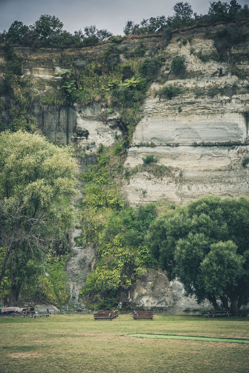 Scenic park benches on green lawn before towering white limestone cliffs with lush green trees and vegetation.