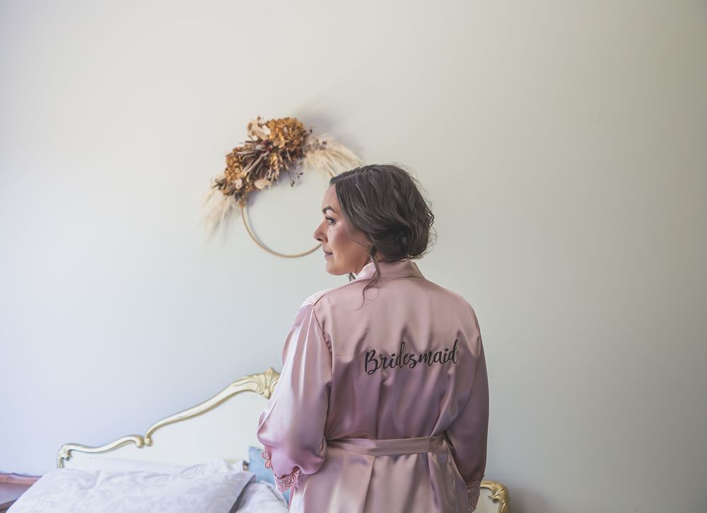A person wearing a pink silk robe sits on a bed near a floral wreath hanging on a white wall.