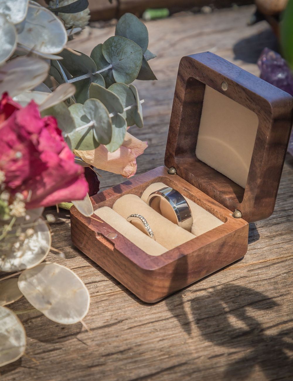 Wedding rings displayed in a wooden box alongside eucalyptus and pink floral decorations on a rustic wooden surface.