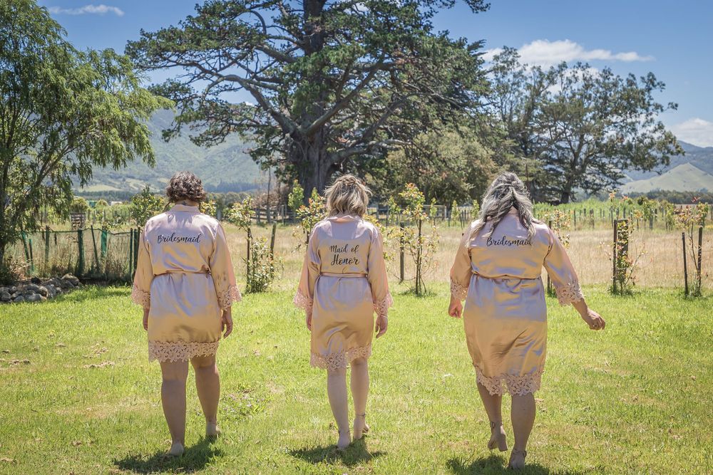 Three people in matching tan bridesmaid dresses walking away from camera on a sunny day in a grassy field.