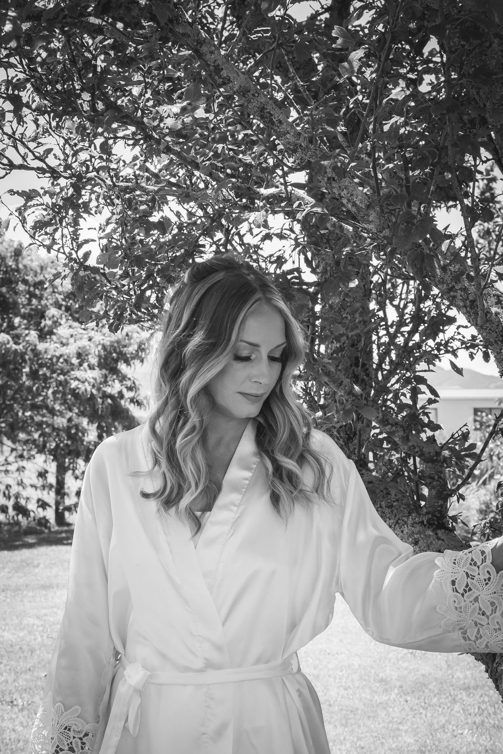 Black and white portrait of a person in a white robe standing outdoors near leafy trees on a sunny day.