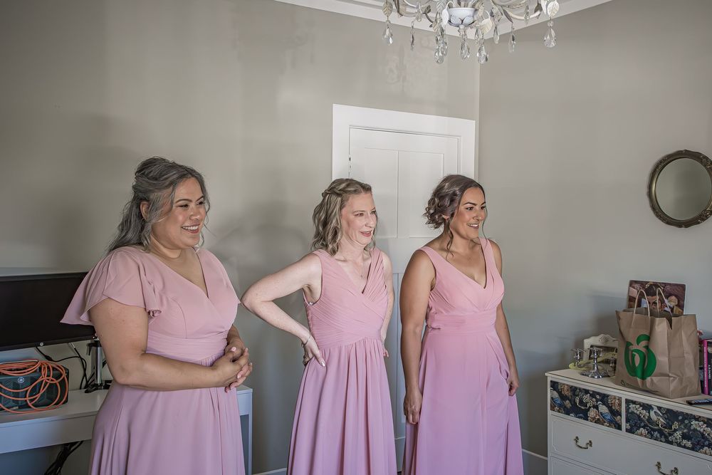 Three people in matching pink dresses stand together in a room with a chandelier and white walls.