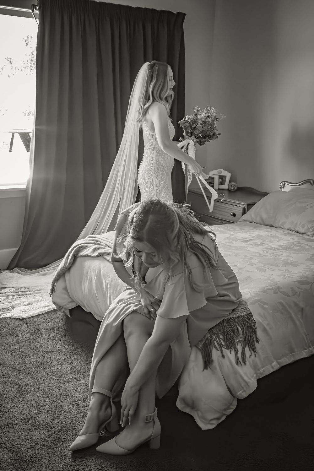 Candid black and white photo of a bride in lace dress standing while someone helps adjust their shoes by the bed.