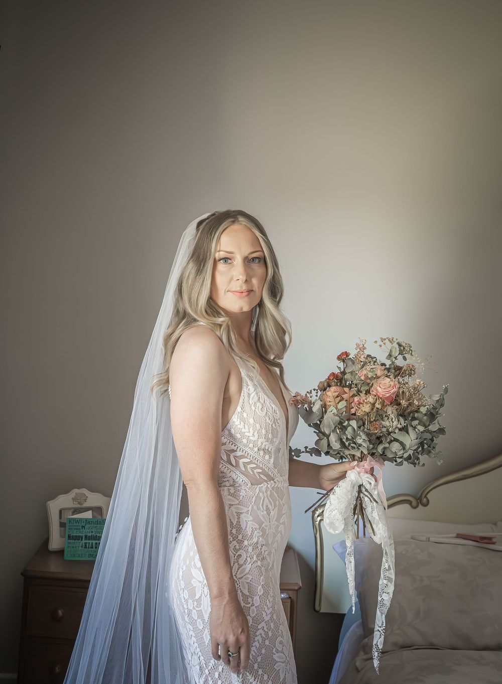 A bride in a lace wedding gown and long veil poses with a soft pink and white floral bouquet against a neutral backdrop.