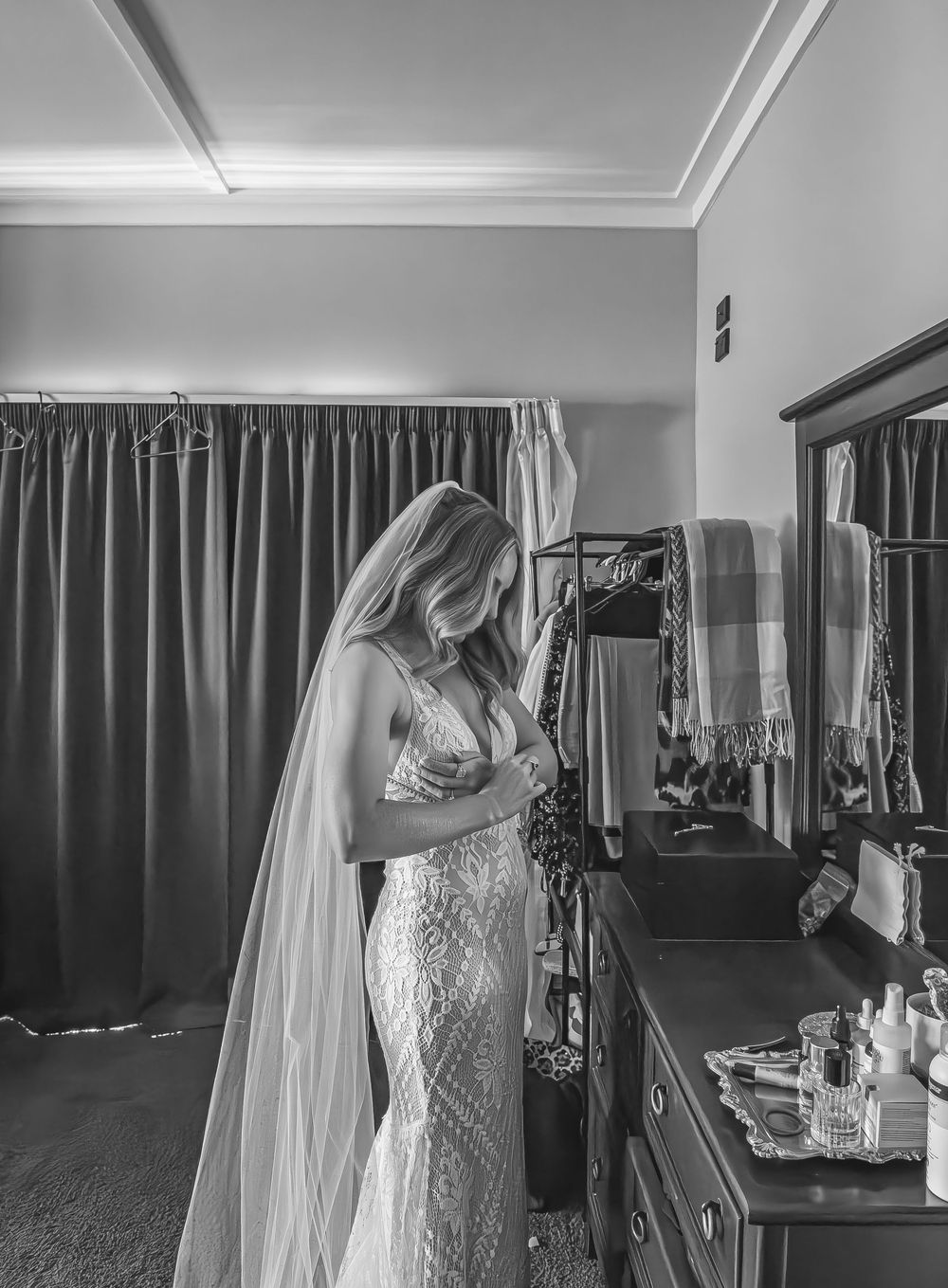 Black and white portrait of a bride in lace wedding gown and veil standing beside a dresser mirror.