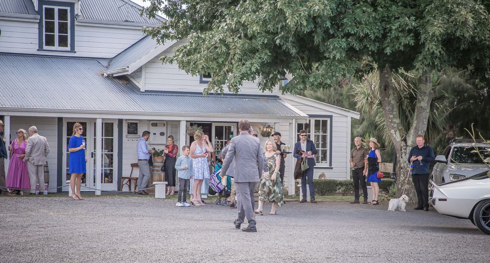 People gather outside a rustic white building with classic architecture during a casual outdoor event.