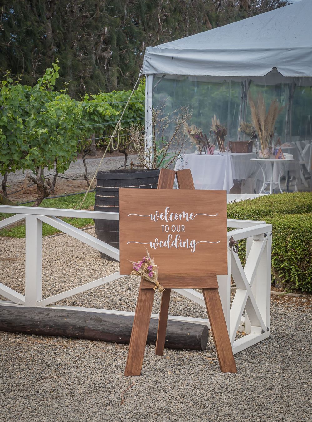 Rustic wooden welcome sign with floral accent stands near a white fence and covered outdoor wedding reception tent.