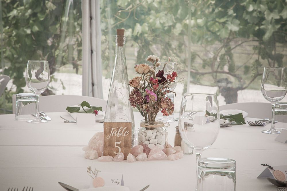 Rustic wedding table centerpiece with dried flowers, glass bottle and table number displayed on white tablecloth.