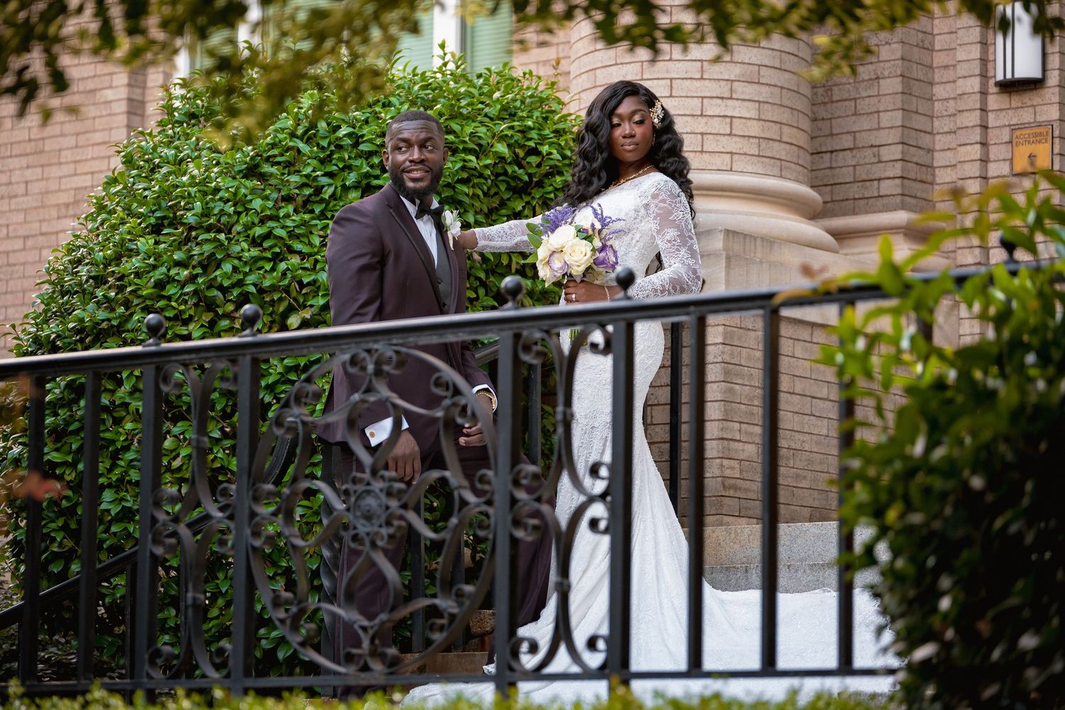Elegant bride and groom posing on ornate iron staircase outside brick building with lush greenery backdrop.