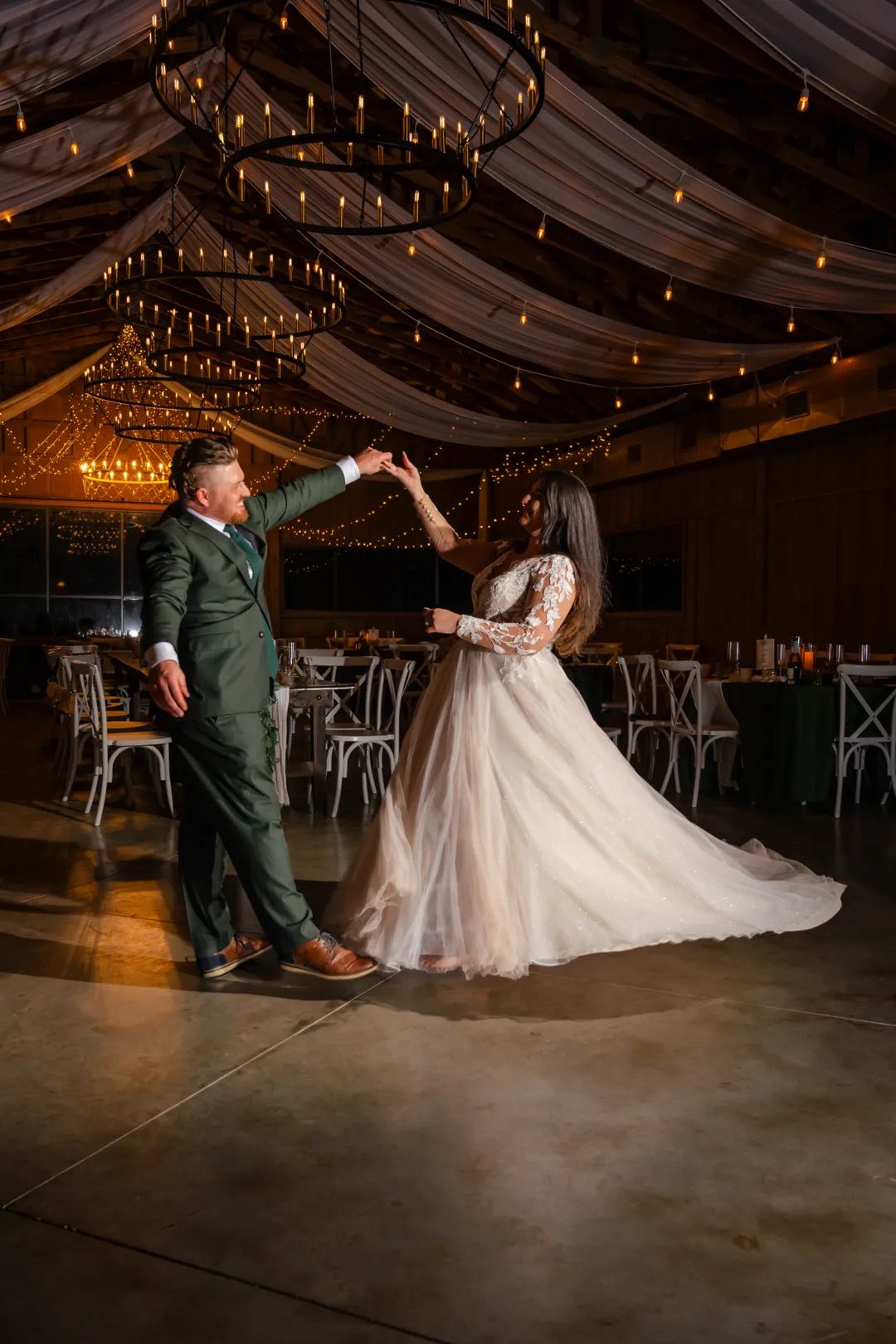 Bride and groom share first dance in elegant barn venue with draped ceilings, string lights, and chandeliers.