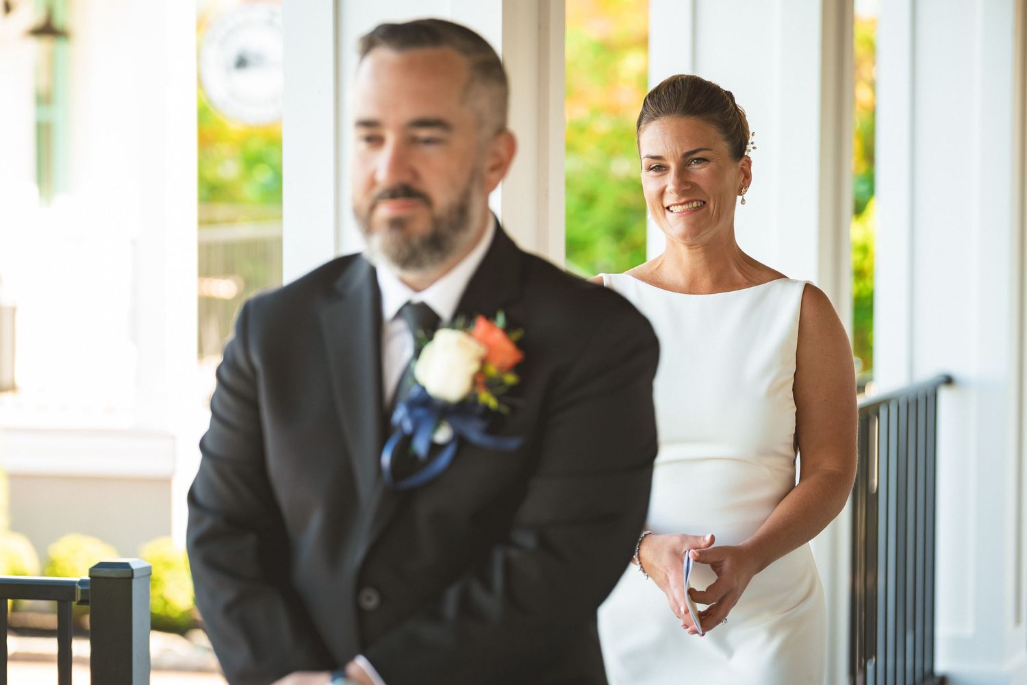 Bride in white gown smiling at groom in black suit with floral boutonniere during wedding first look ceremony.