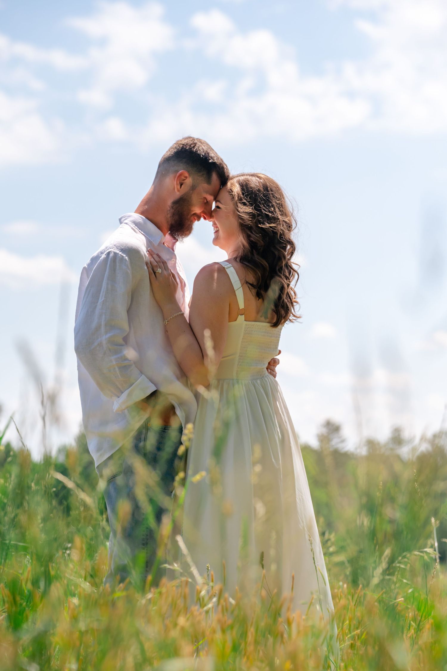 Romantic couple embracing in a sunny green field, foreheads touching, wearing white outfits under a bright blue sky.