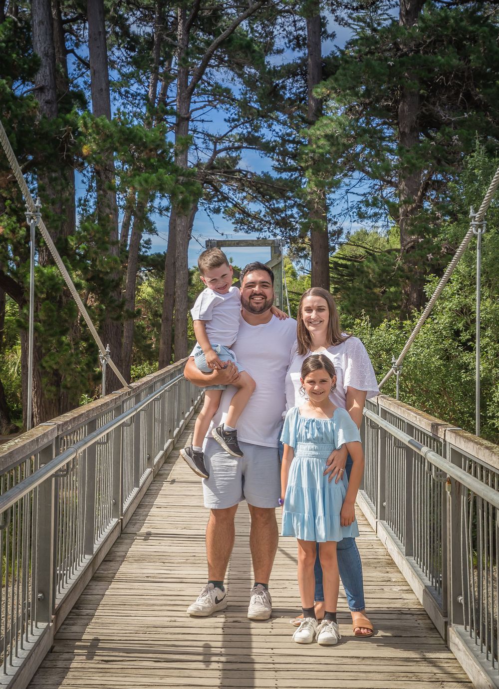 A family poses together on a wooden bridge surrounded by tall trees in a natural outdoor setting.